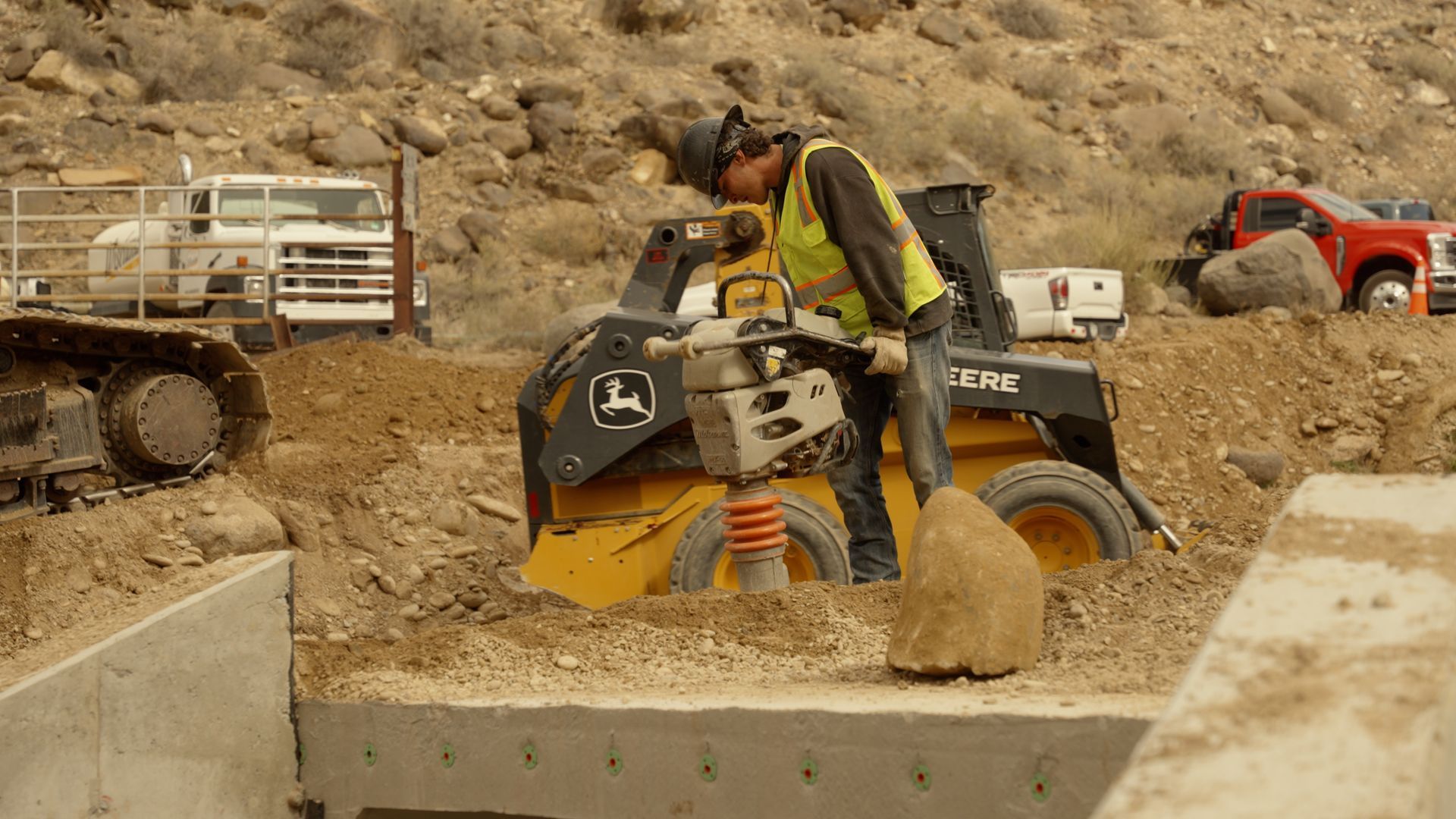 Worker operating a jackhammer in a dusty construction site with vehicles in the background