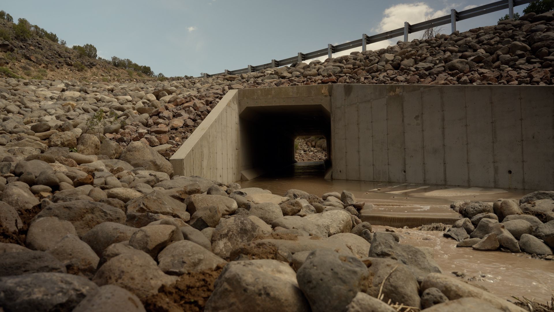 Concrete drainage tunnel opening beneath a rocky embankment, with boulders scattered in the foreground.