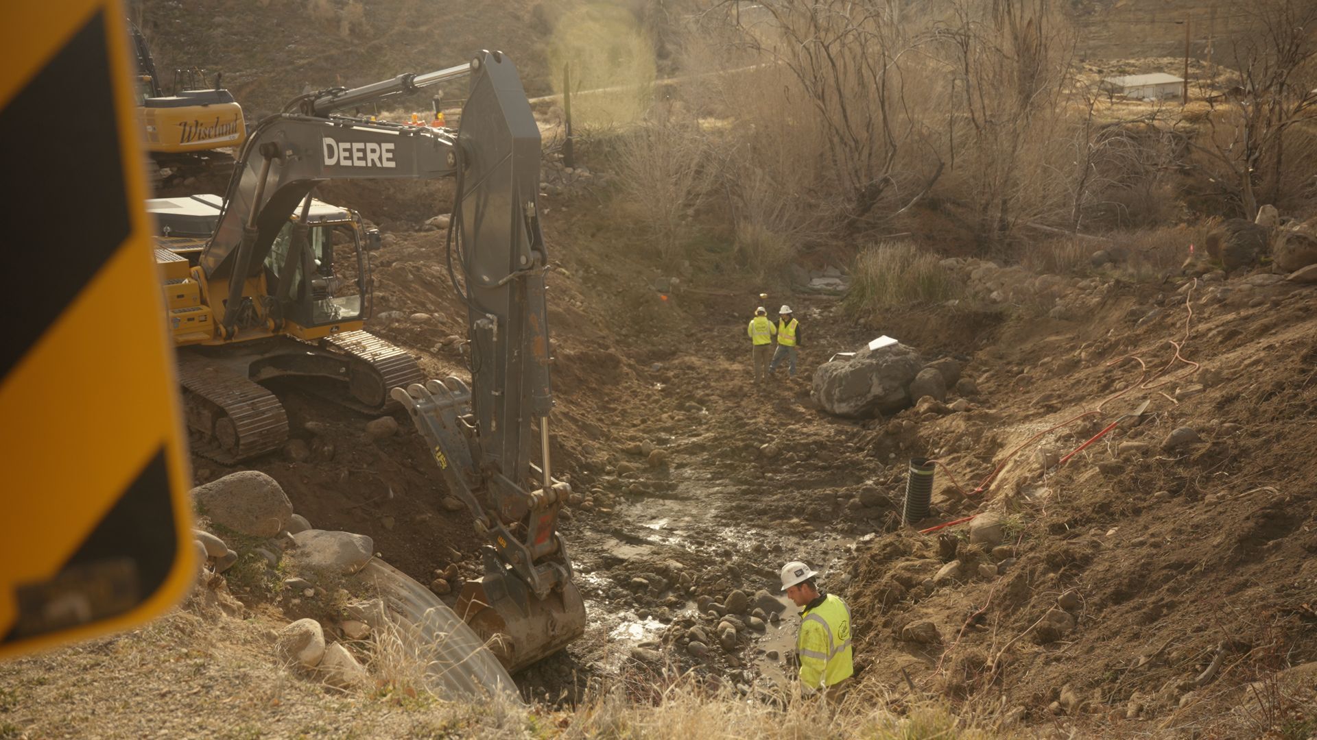 Construction workers in neon vests in a muddy excavation site with an excavator and large rocks.