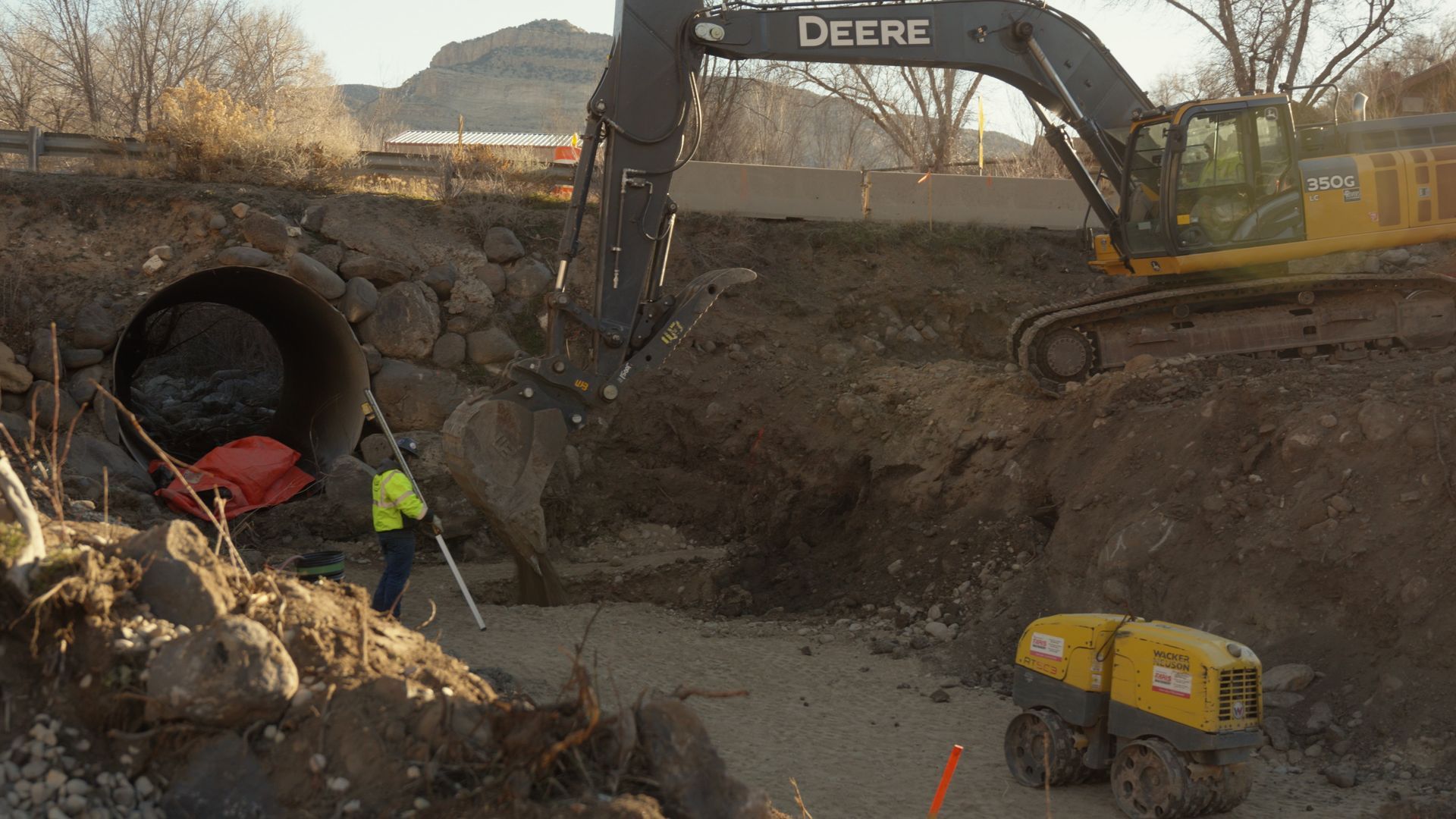 Excavator and workers repairing a collapsed culvert near a large pipe under a dirt road.