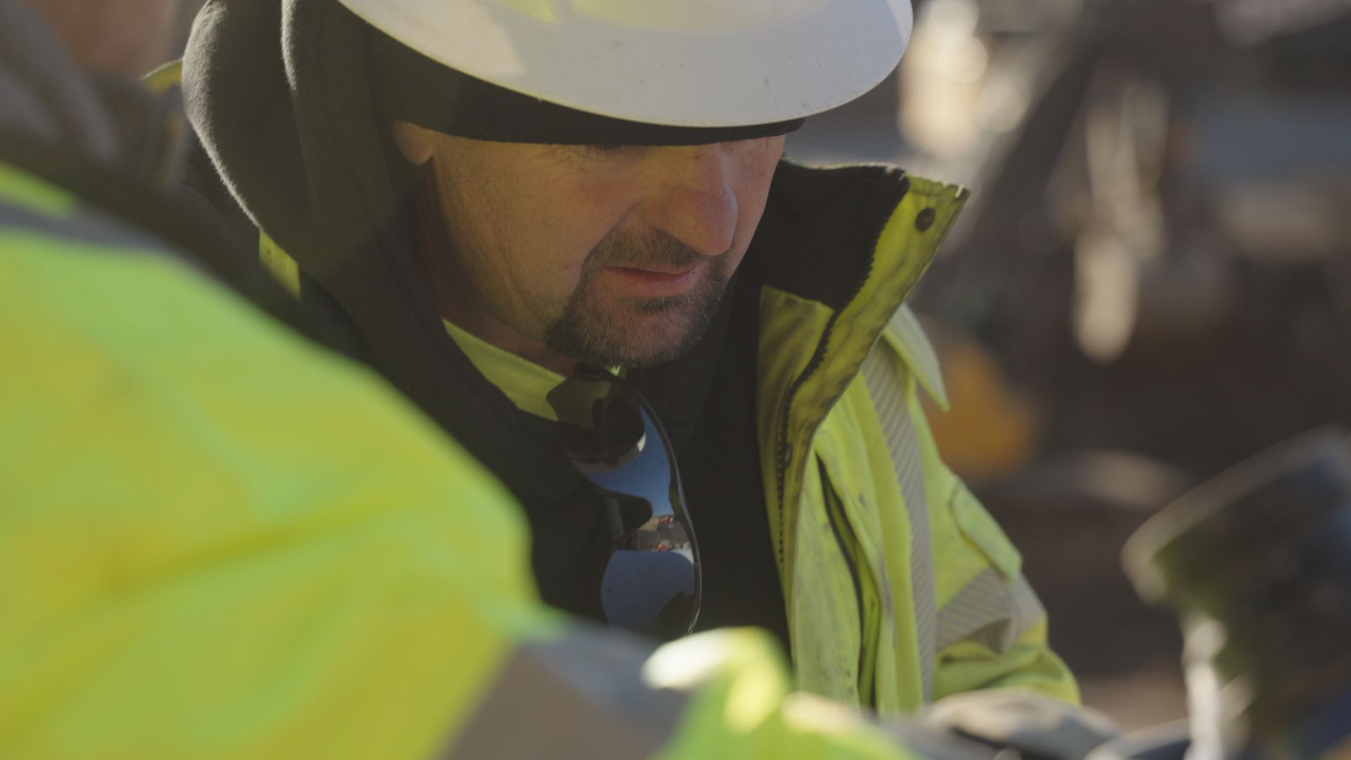 Worker in white hard hat and yellow safety jacket, looking down at a construction site.