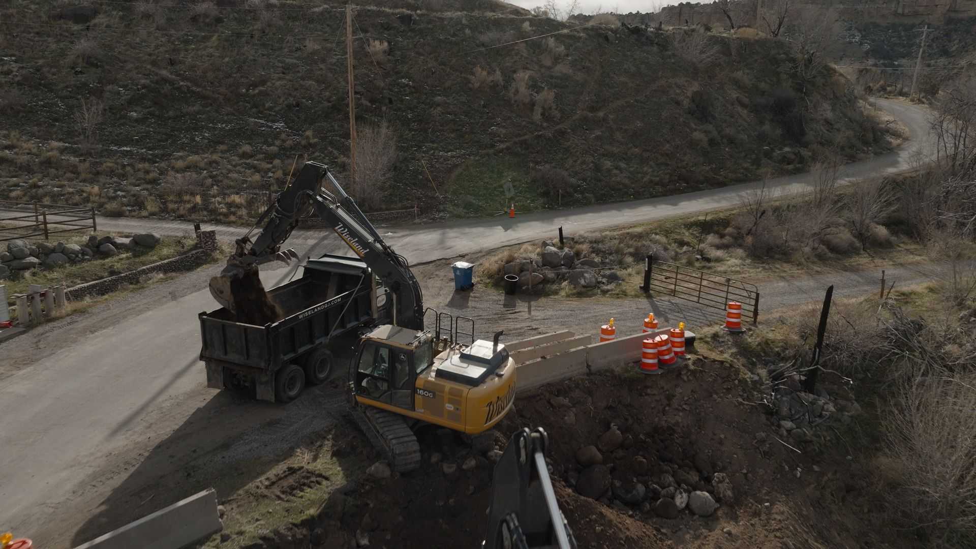 Excavator loading a dump truck at a dirt construction site beside a winding road
