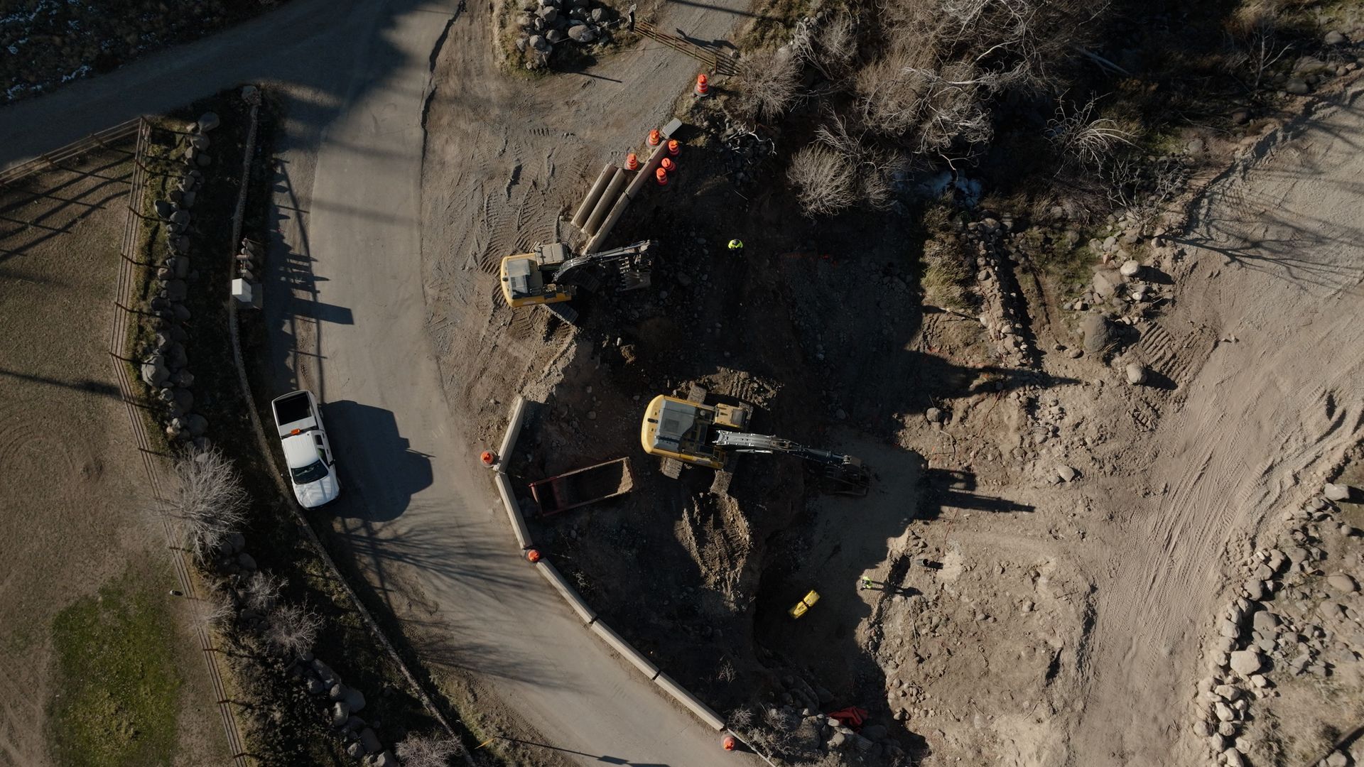 Aerial view of a construction site beside a curved road, with excavators and dirt piles.
