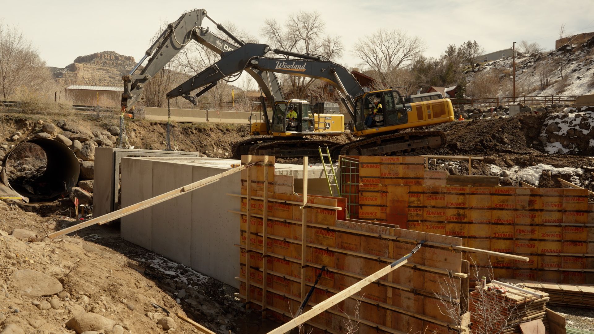 Excavator working at a construction site beside a large concrete trench and wooden retaining wall.
