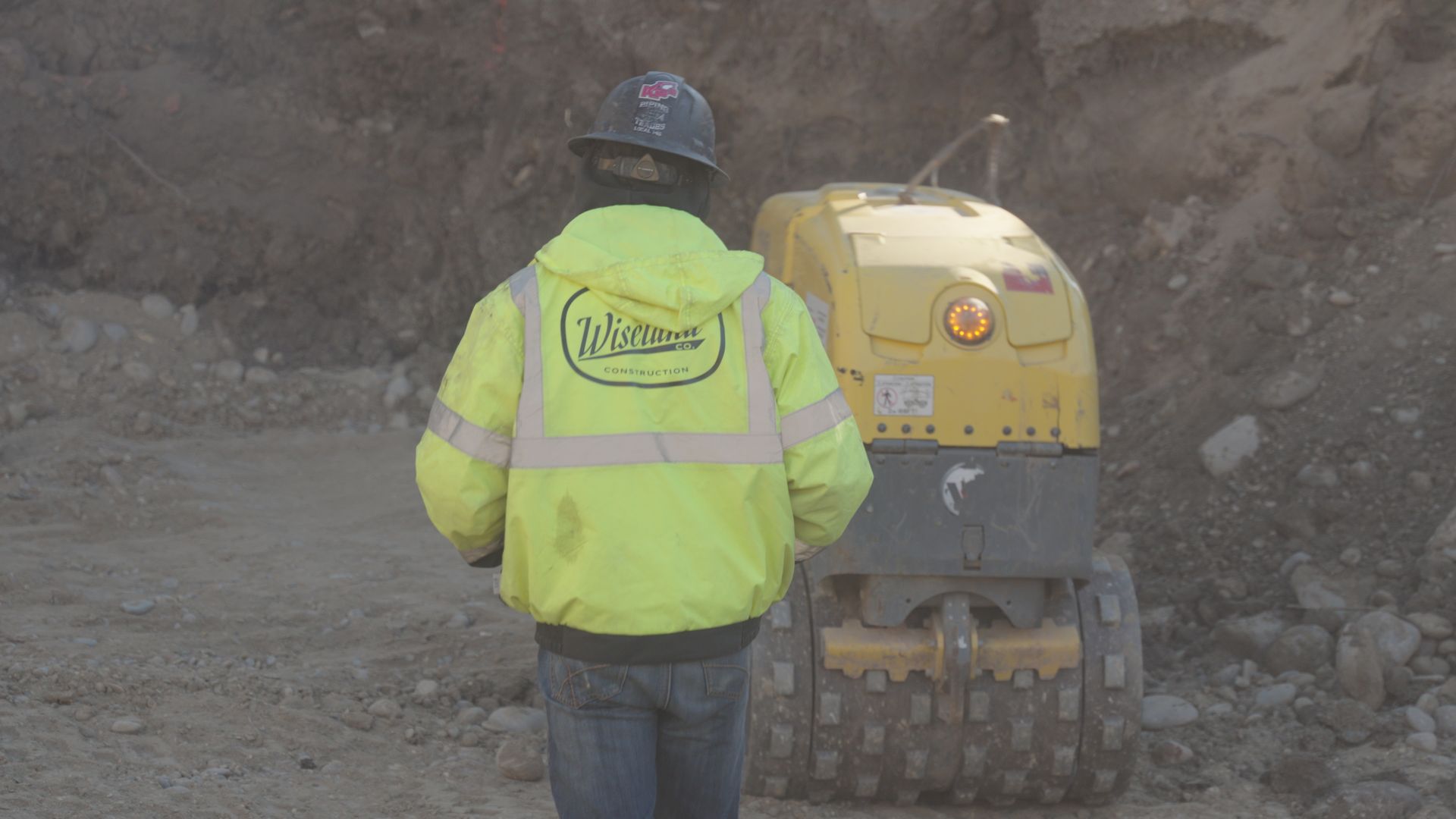 Worker in a neon jacket and hard hat stands beside a small yellow construction machine in a rocky excavation site.