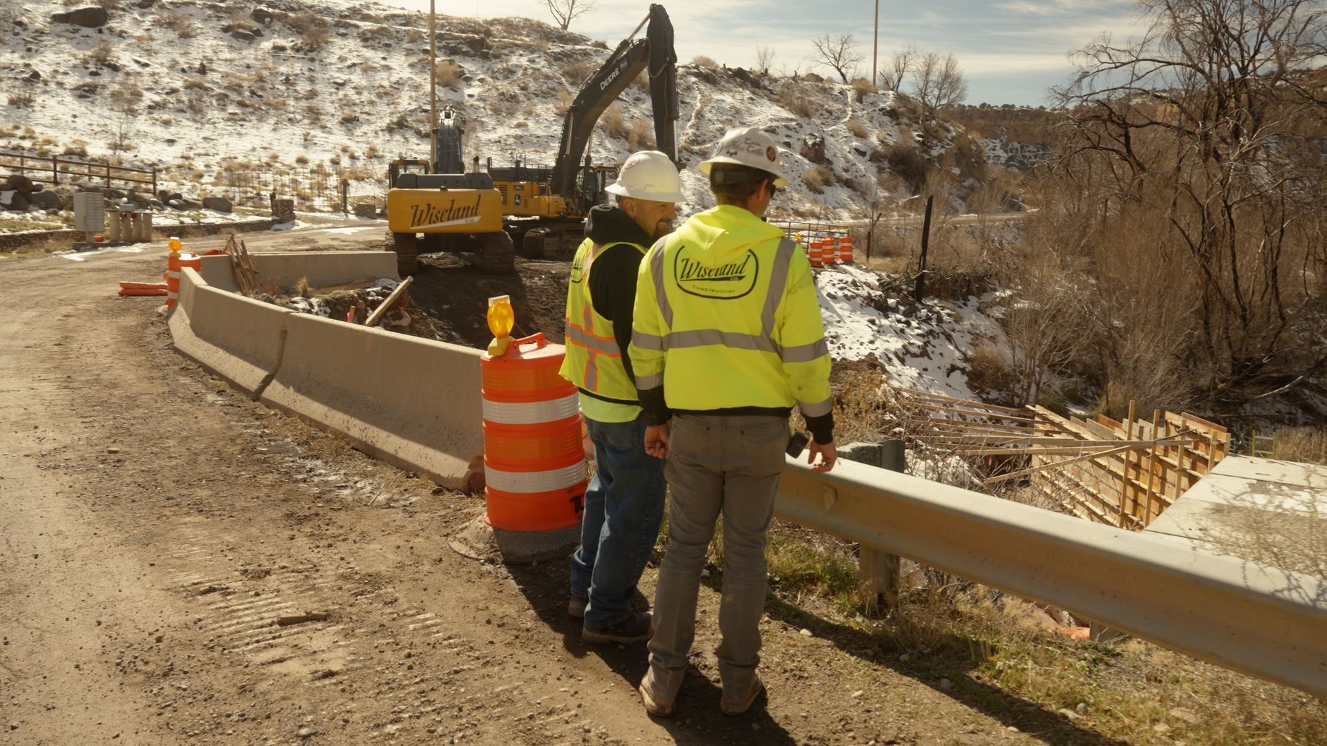 Construction workers in yellow vests near a roadwork barrier and excavator on a snowy roadside