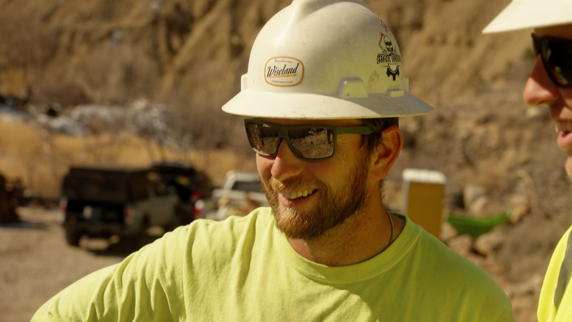 Construction worker in white hard hat and glasses smiling outdoors in a rocky, dusty site