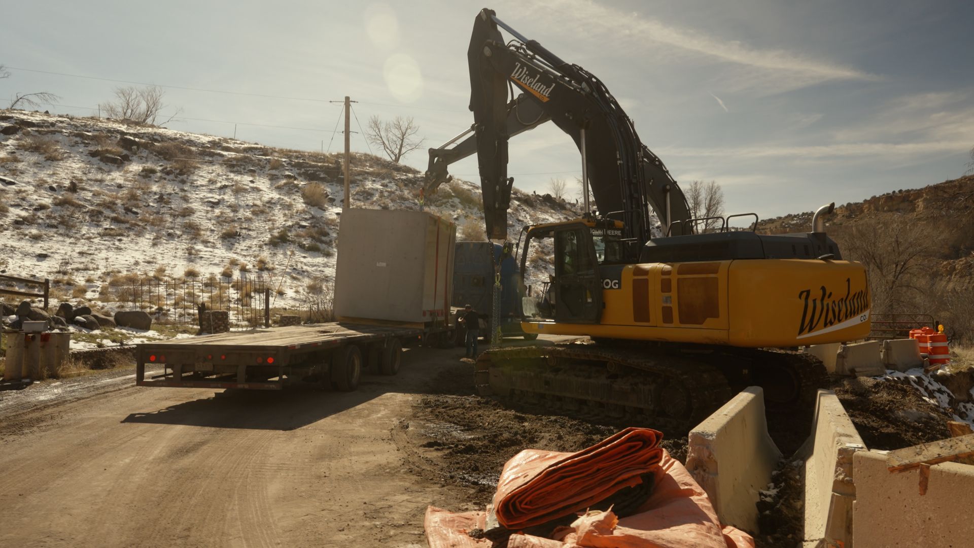 Excavator loading a flatbed truck at a dusty construction site beside a snowy hillside.