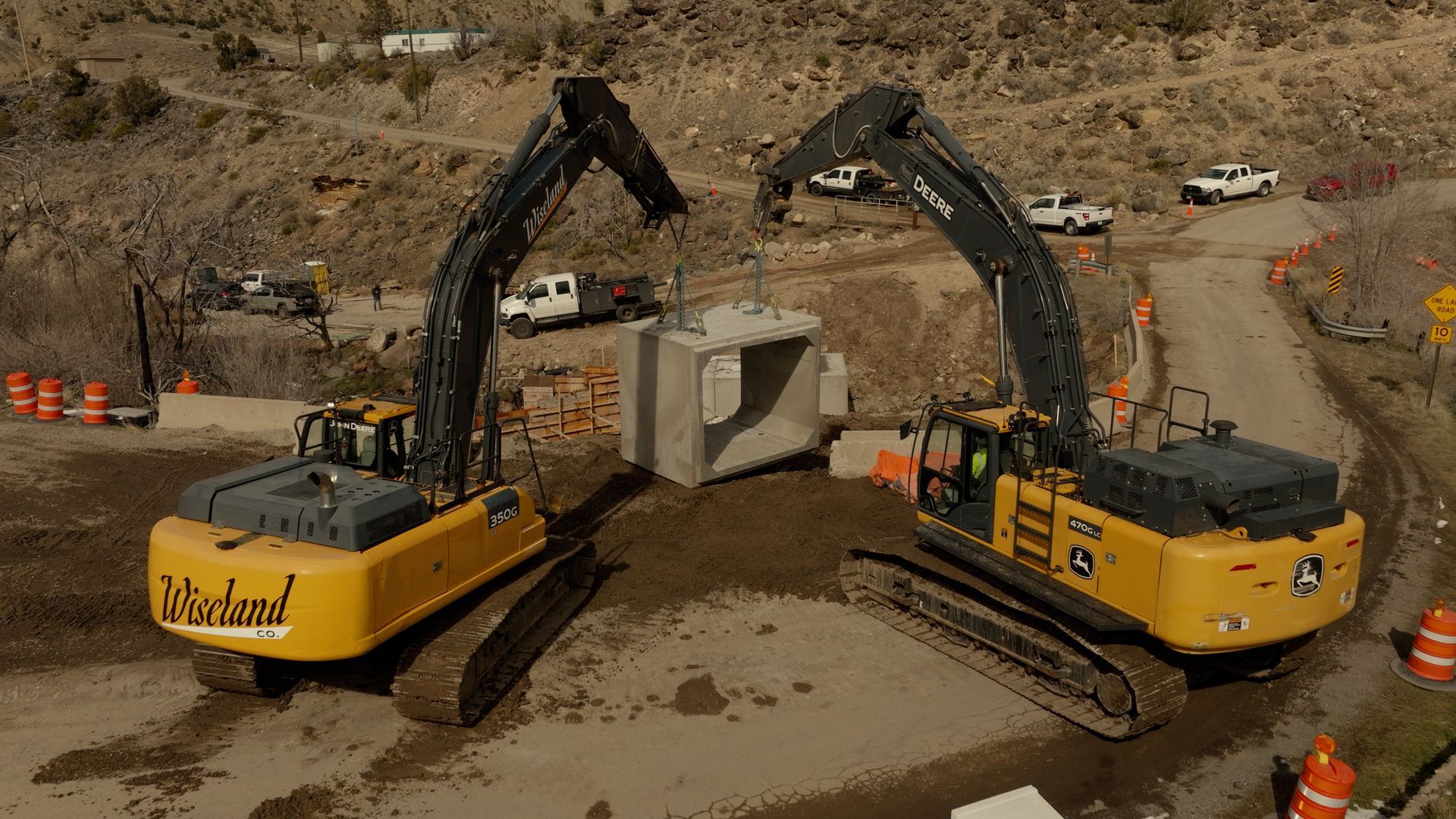 Two yellow excavators lift a concrete block at a construction site.