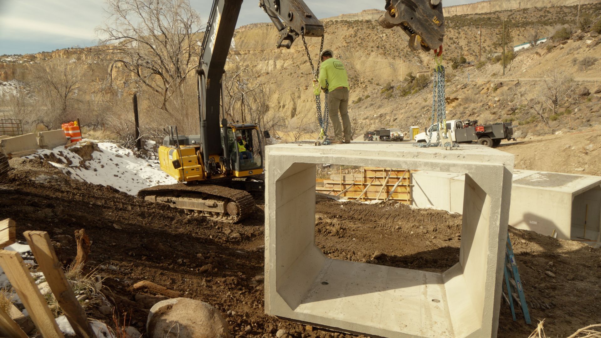 Construction site with excavator, workers, and concrete drainage blocks in a dirt trench