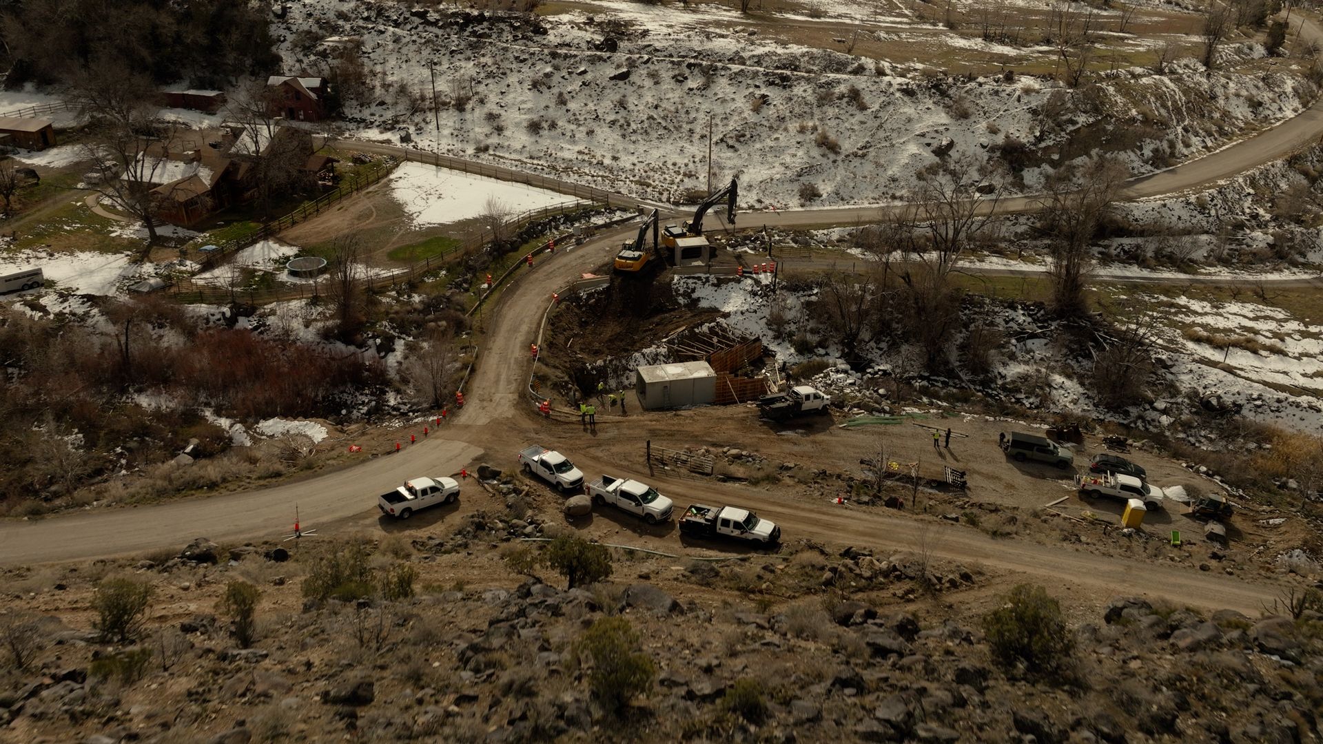 A dusty road winds through a rocky hillside, with several parked vehicles and a few people near the bend.