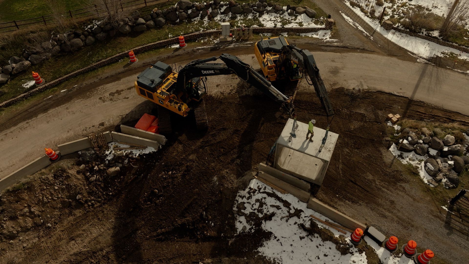 Excavator loads a concrete block at a muddy roadside construction site with orange cones nearby
