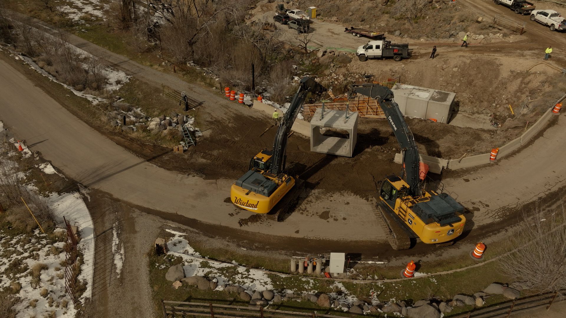Construction excavators shaping a circular dirt basin at a worksite, with orange cones and trucks nearby