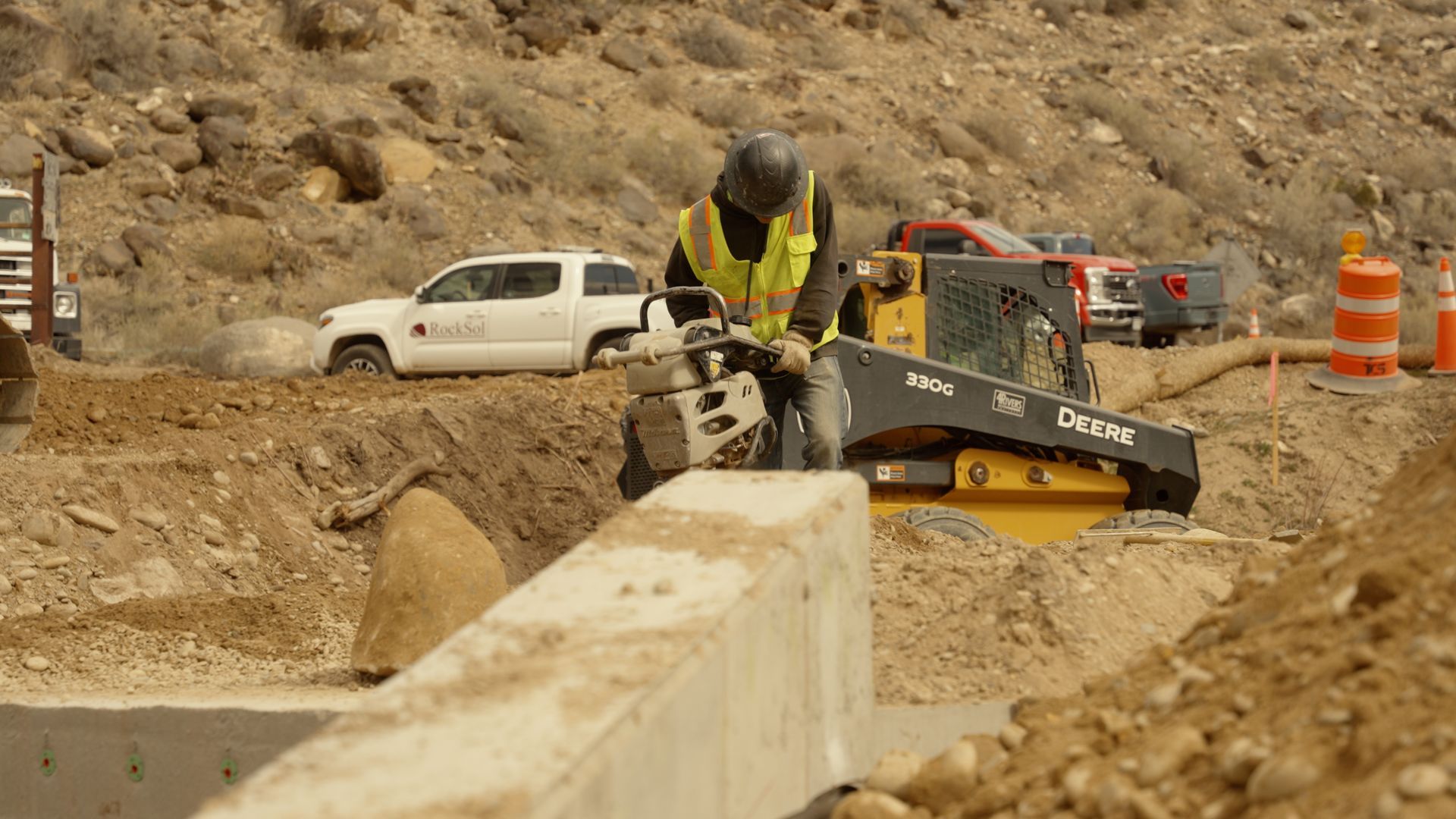 Construction worker operates a jackhammer beside a concrete barrier at a dusty roadwork site.