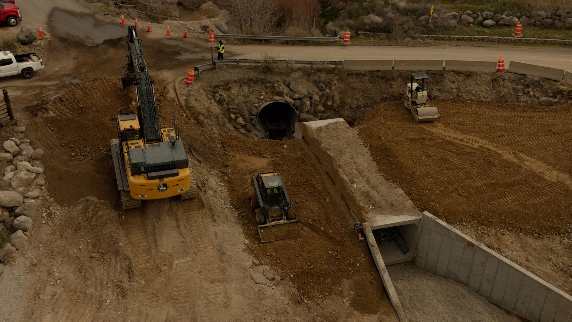 Construction site with excavator, dump truck, and workers near a tunnel entrance and concrete trench.