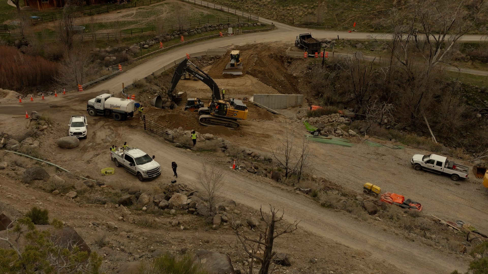 Construction site with excavators, dump trucks, and white vehicles on a dirt road near water.