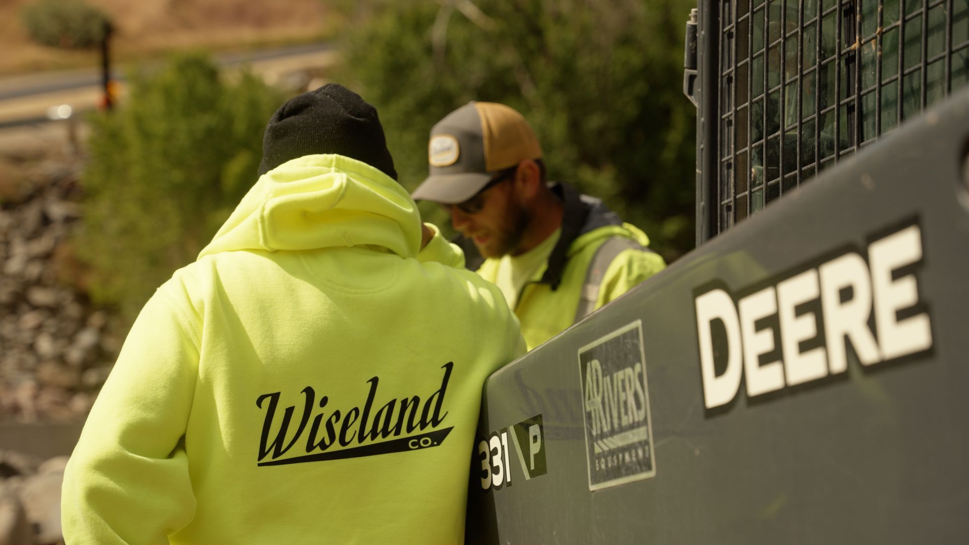 Two workers in neon jackets stand beside a John Deere machine outdoors.