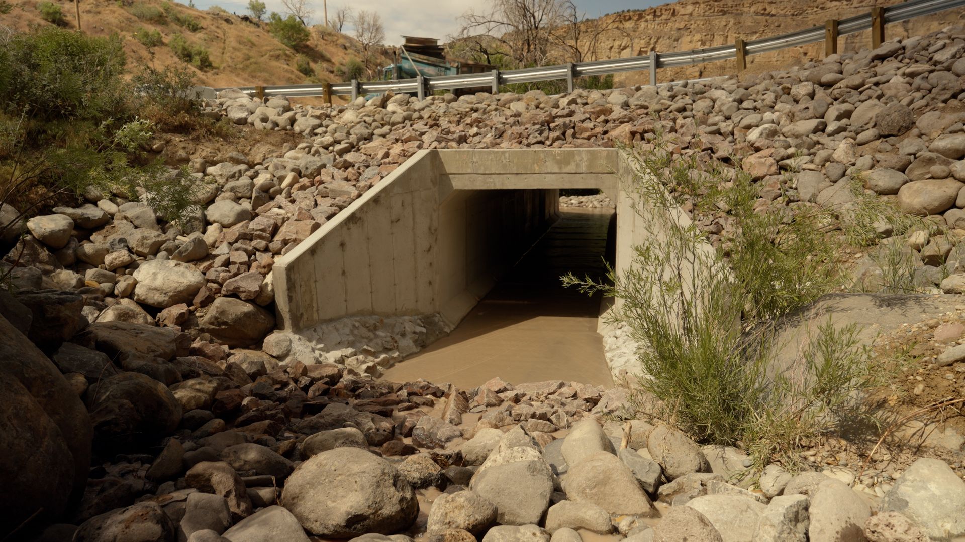 Concrete drainage culvert under a road embankment in a rocky, dry wash with sparse shrubs