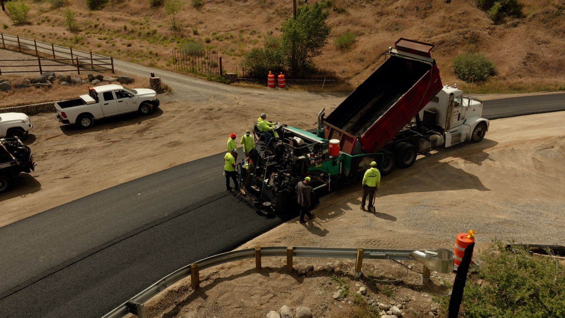 Workers surround a dump truck on a curved mountain road, with emergency vehicles nearby.