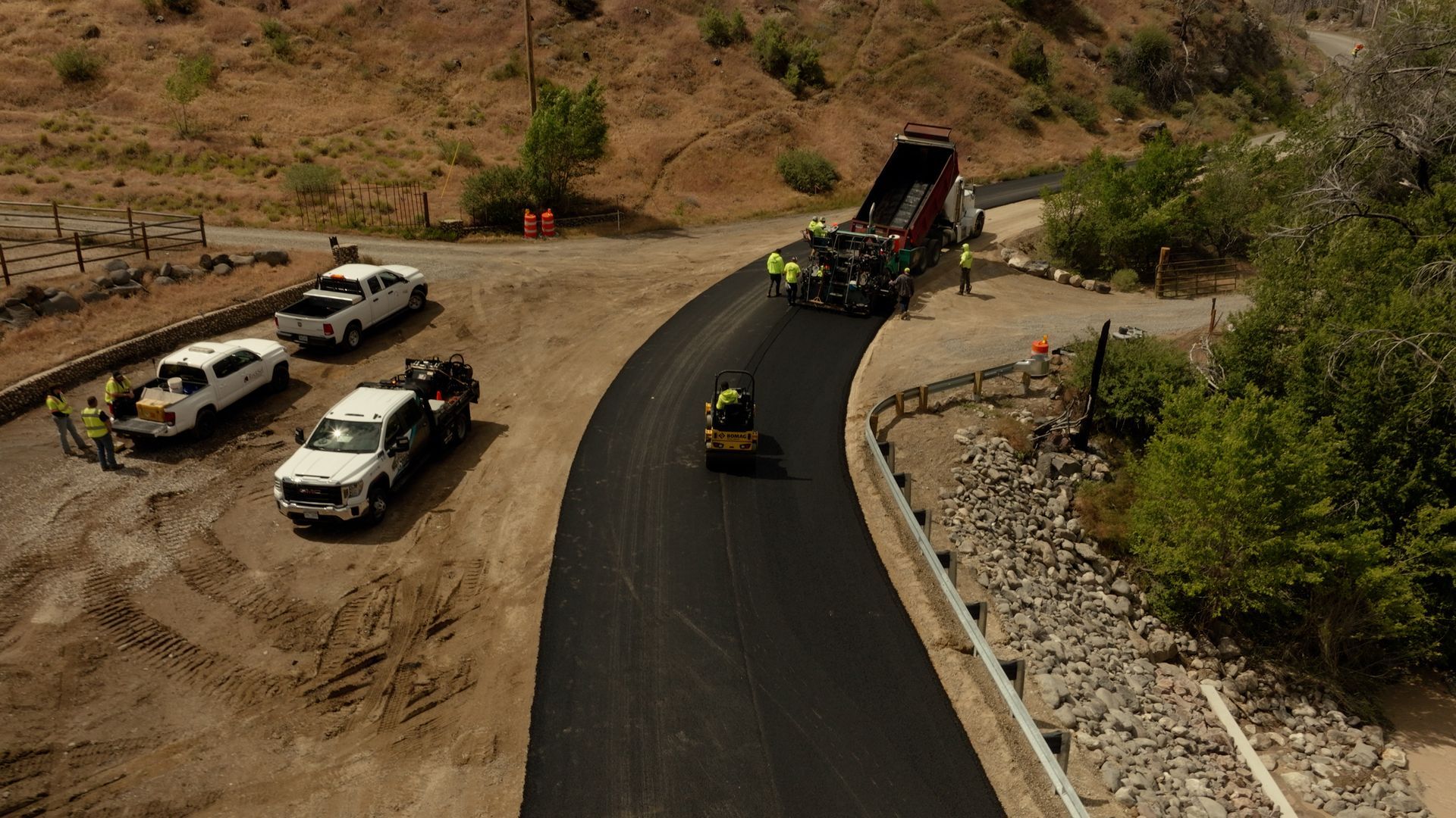 Road construction on a curving hill road with trucks, equipment, and workers near the asphalt.