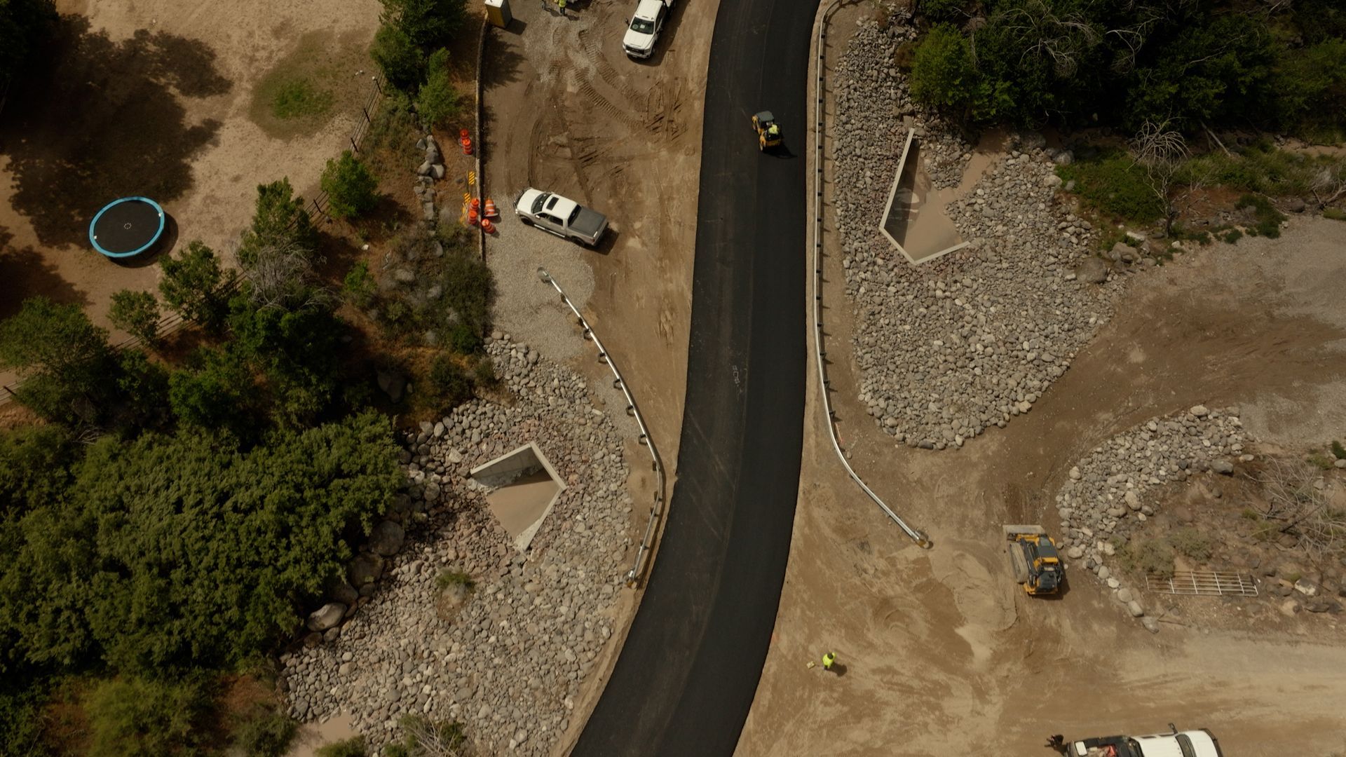 Aerial view of a winding road beside dirt lots, rocks, trees, and a few parked cars.