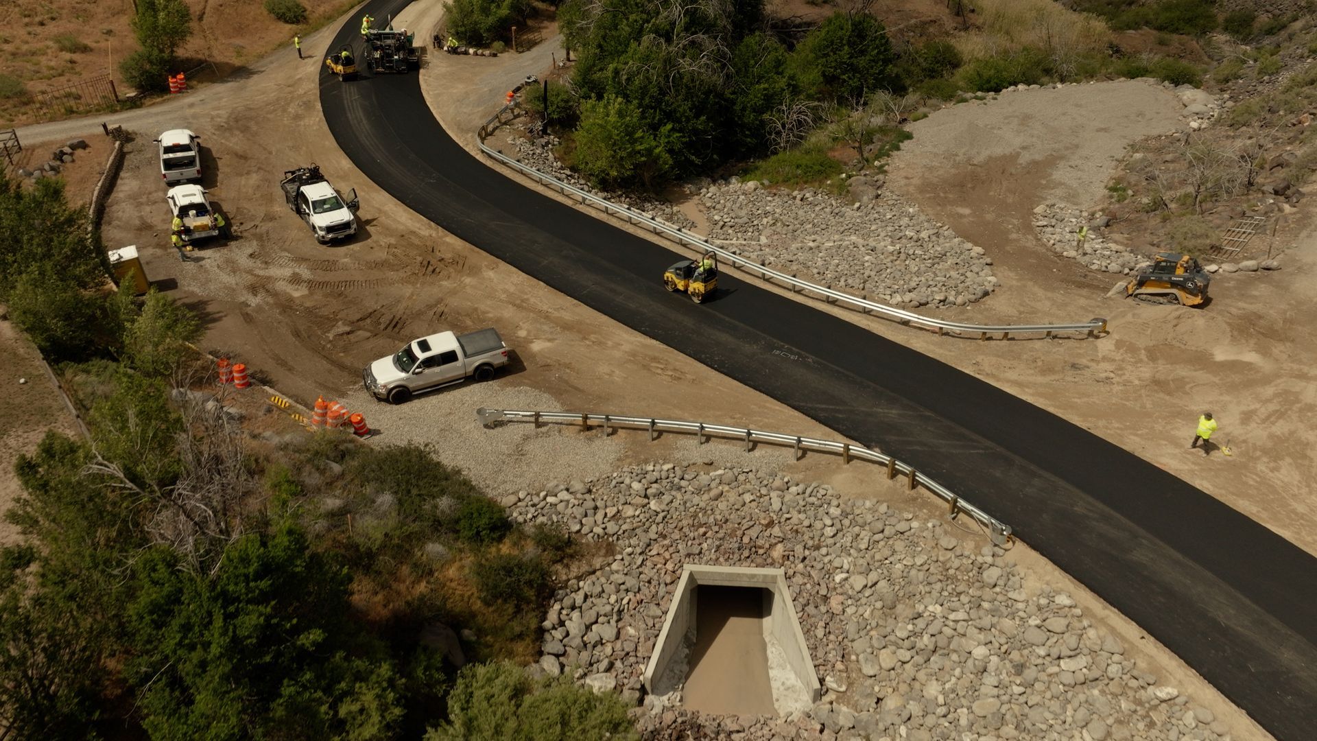 Aerial view of road construction with workers, white trucks, and a new curving asphalt road on rocky terrain