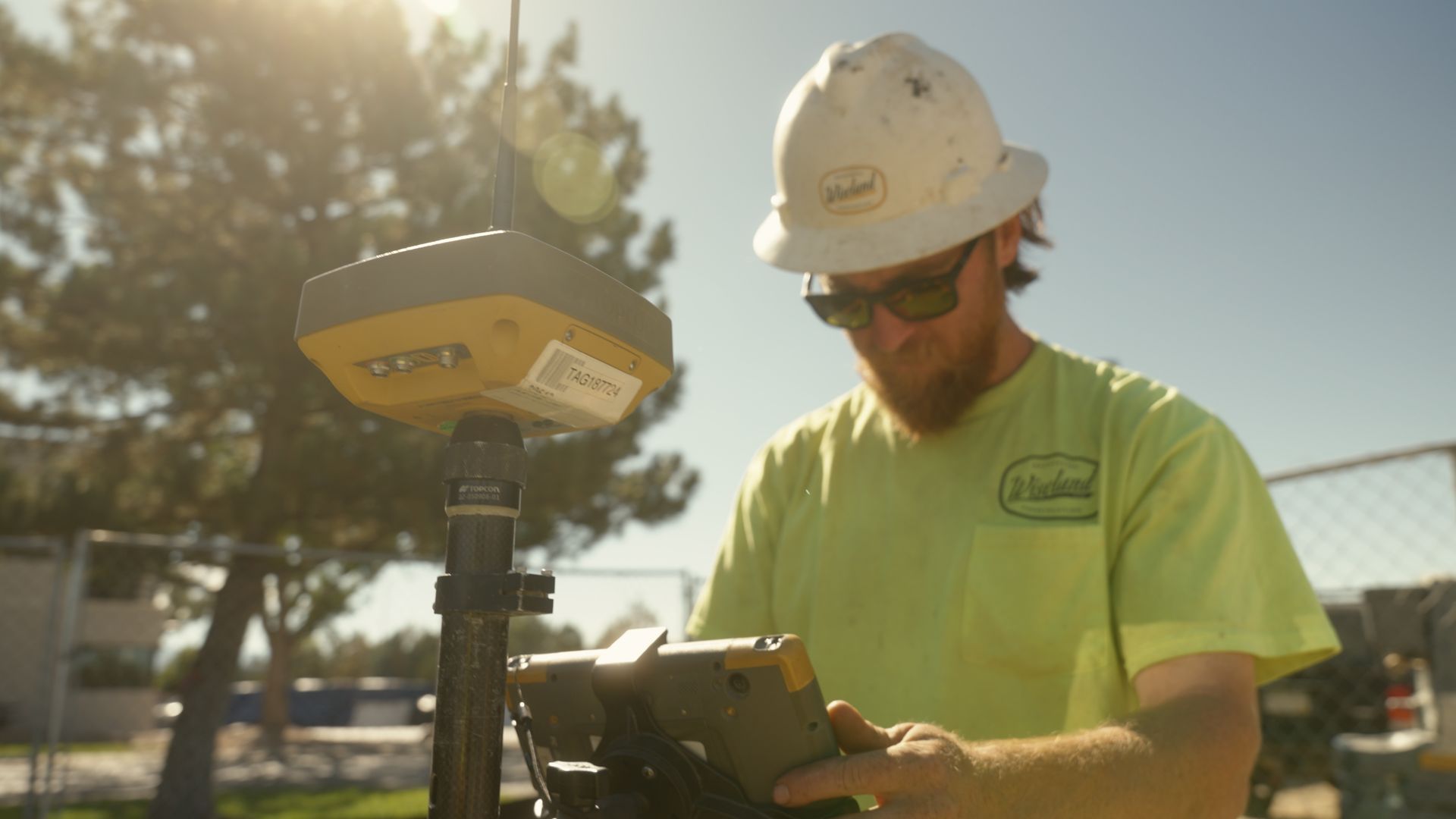 Worker in white hard hat and neon shirt using a tablet beside surveying equipment outdoors