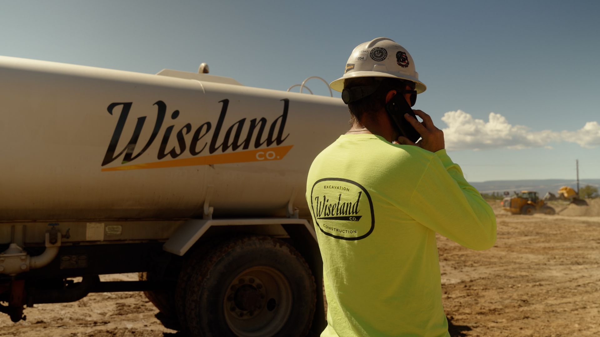Worker in neon shirt and helmet stands beside a Wiseland tanker truck in a dry desert area.