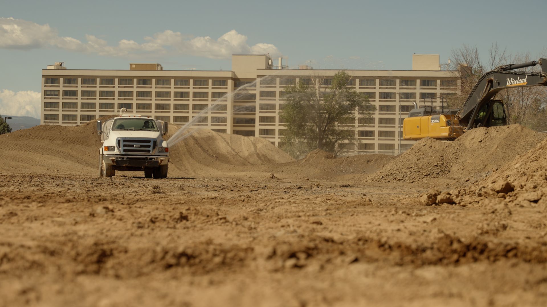 Construction site with a white truck, excavator, and dirt piles in front of a large building.