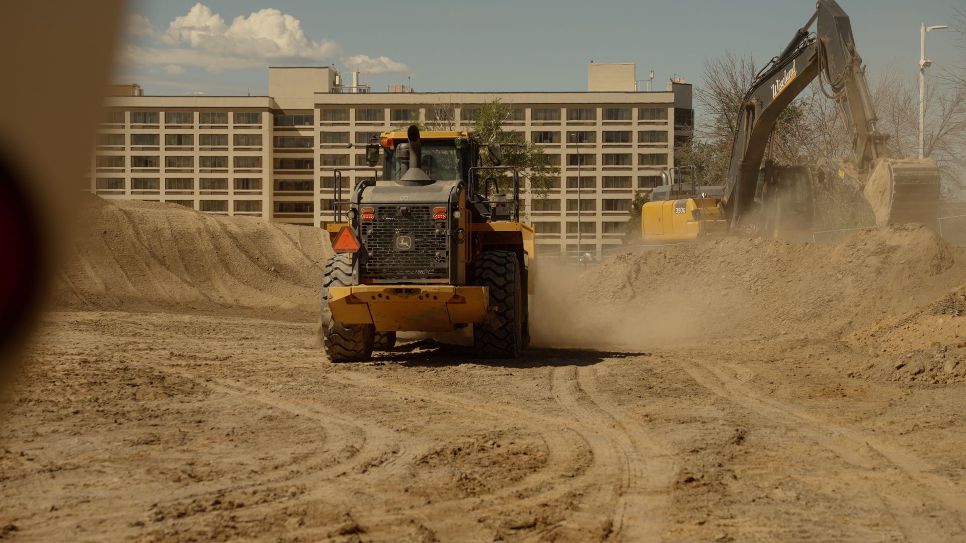 Construction vehicles clearing a dusty site in front of a large building.