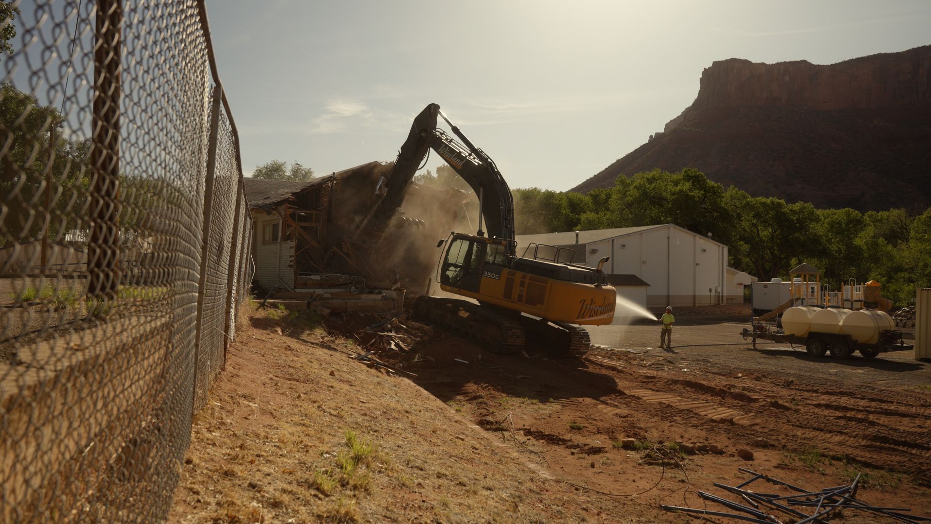 Excavator loading dirt at a construction site beside a chain-link fence and mountain backdrop