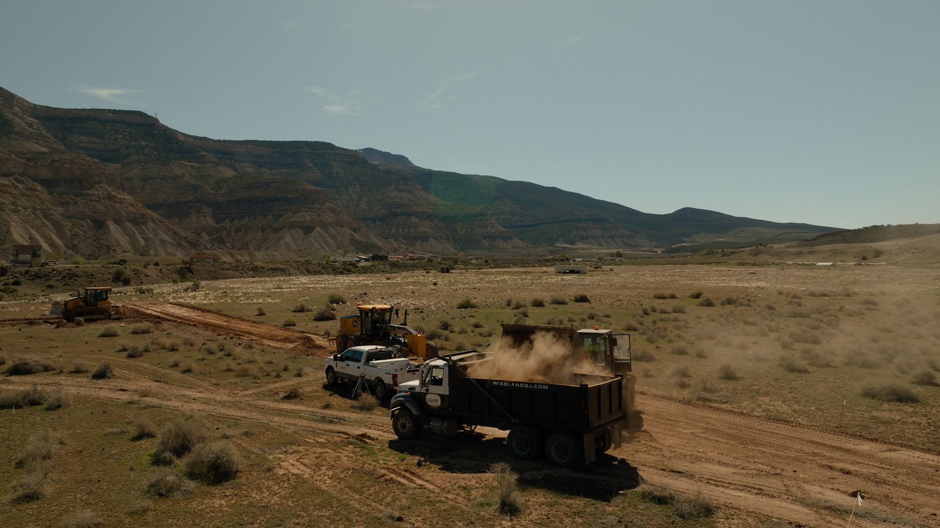 Two construction trucks on a dusty dirt road in a dry, hilly landscape