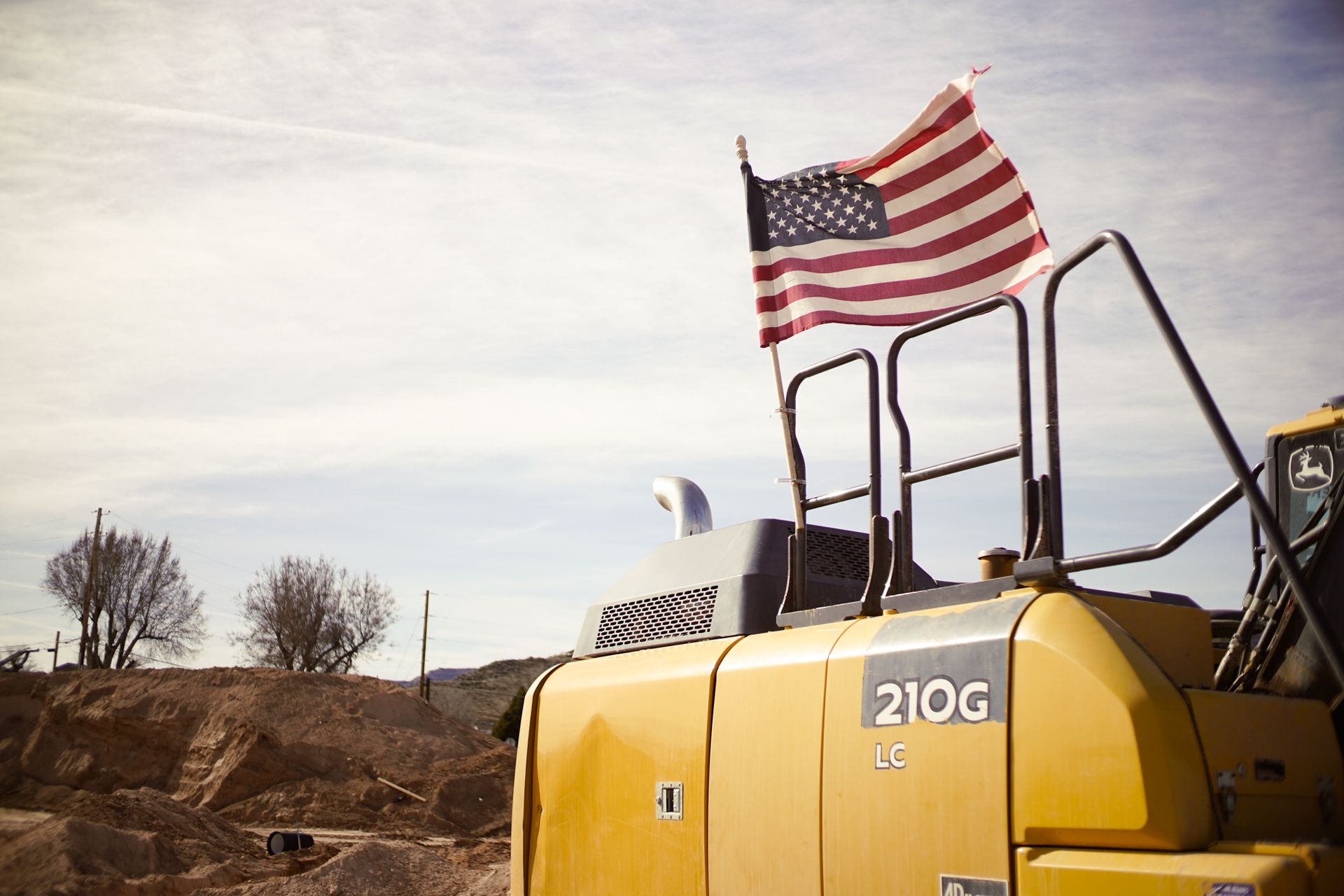 Yellow 210G construction vehicle with an American flag outdoors under a cloudy sky