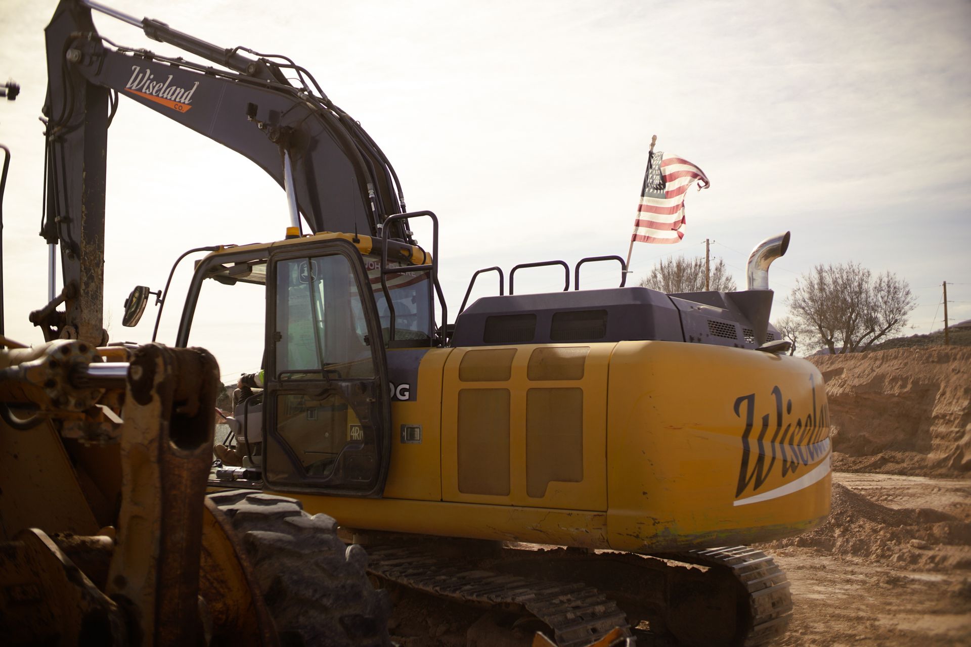 Yellow excavator at a construction site with a raised arm and American flag on top