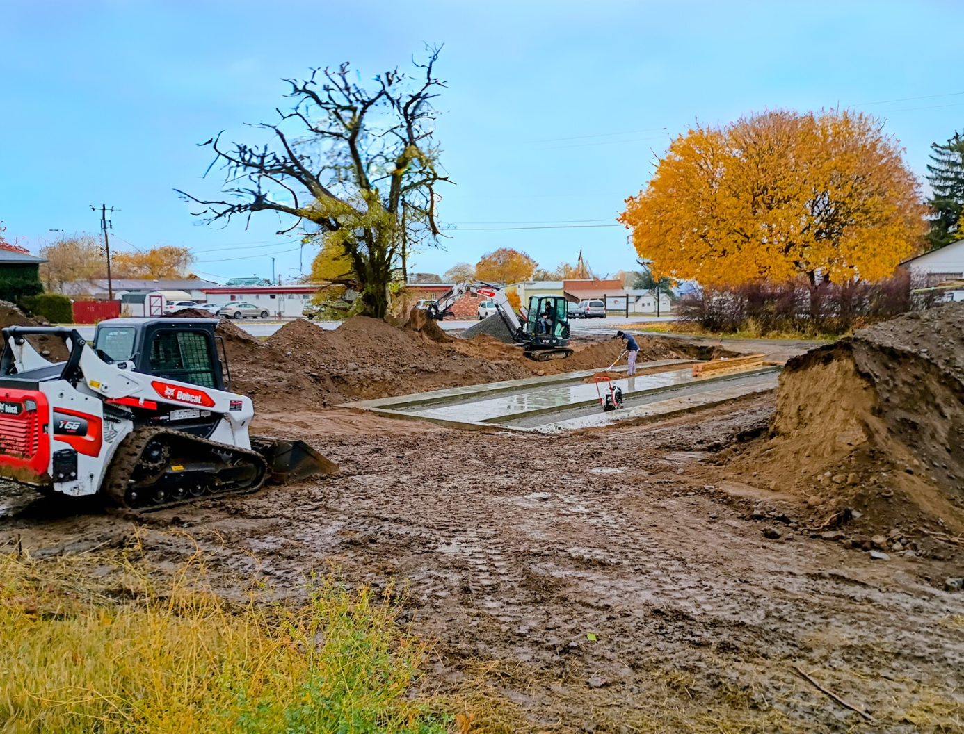 Mud-covered construction site with a skid steer, dirt piles, and autumn trees by a water channel