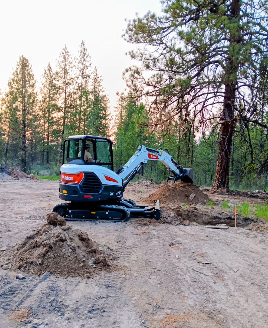 Small excavator clearing dirt beside a forest trail at dusk