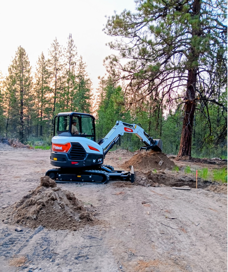 Small excavator digging dirt at a wooded construction site at dusk