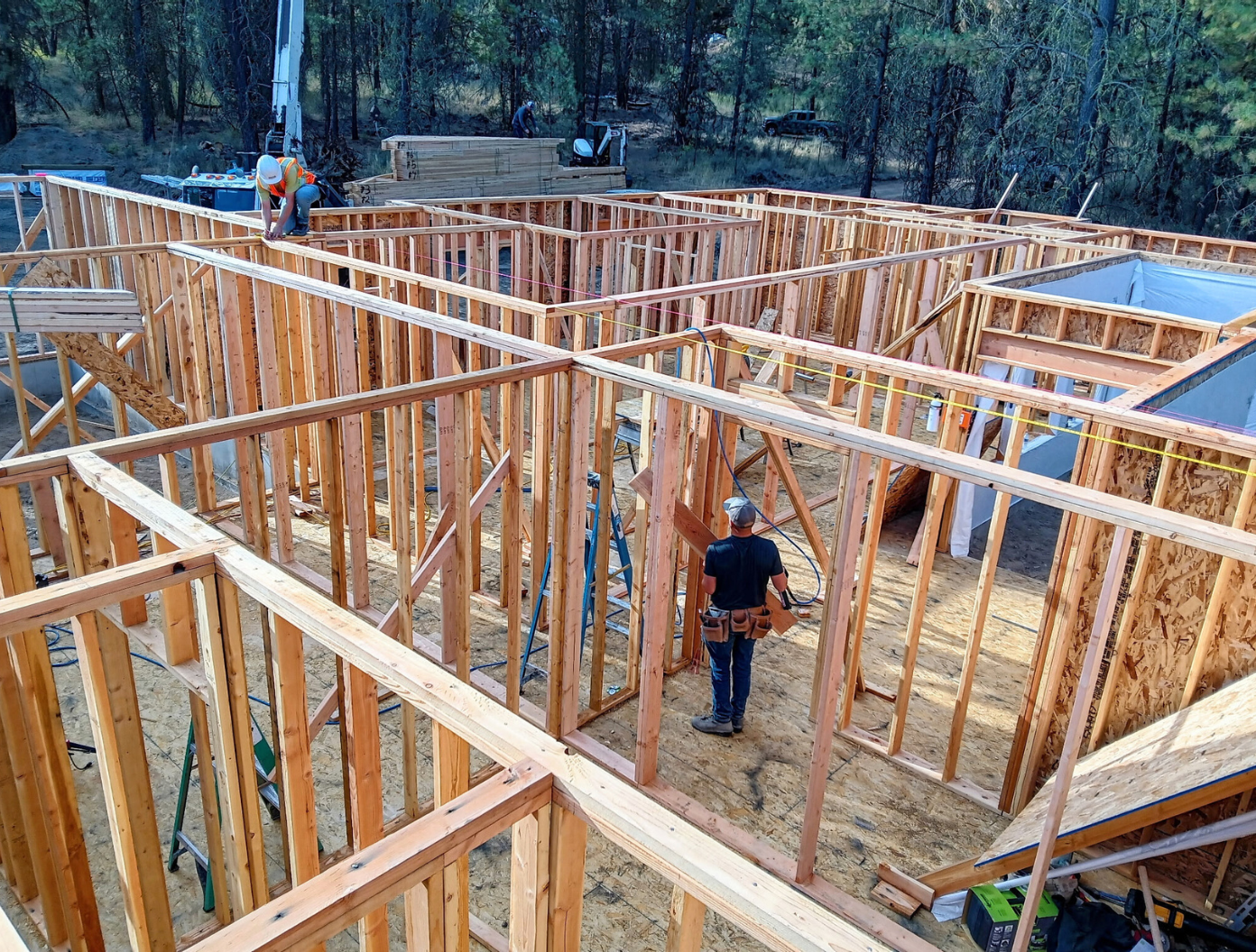 Wood-framed house under construction in a wooded area, with one worker standing inside the framing.