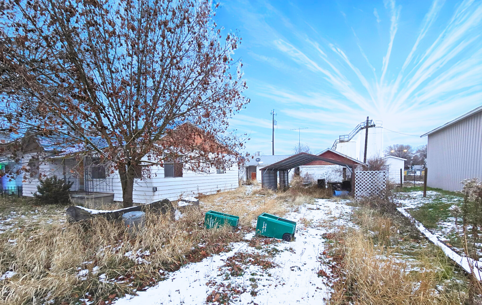 Snowy rural yard with a bare tree, green bins, and small houses under a bright blue sky