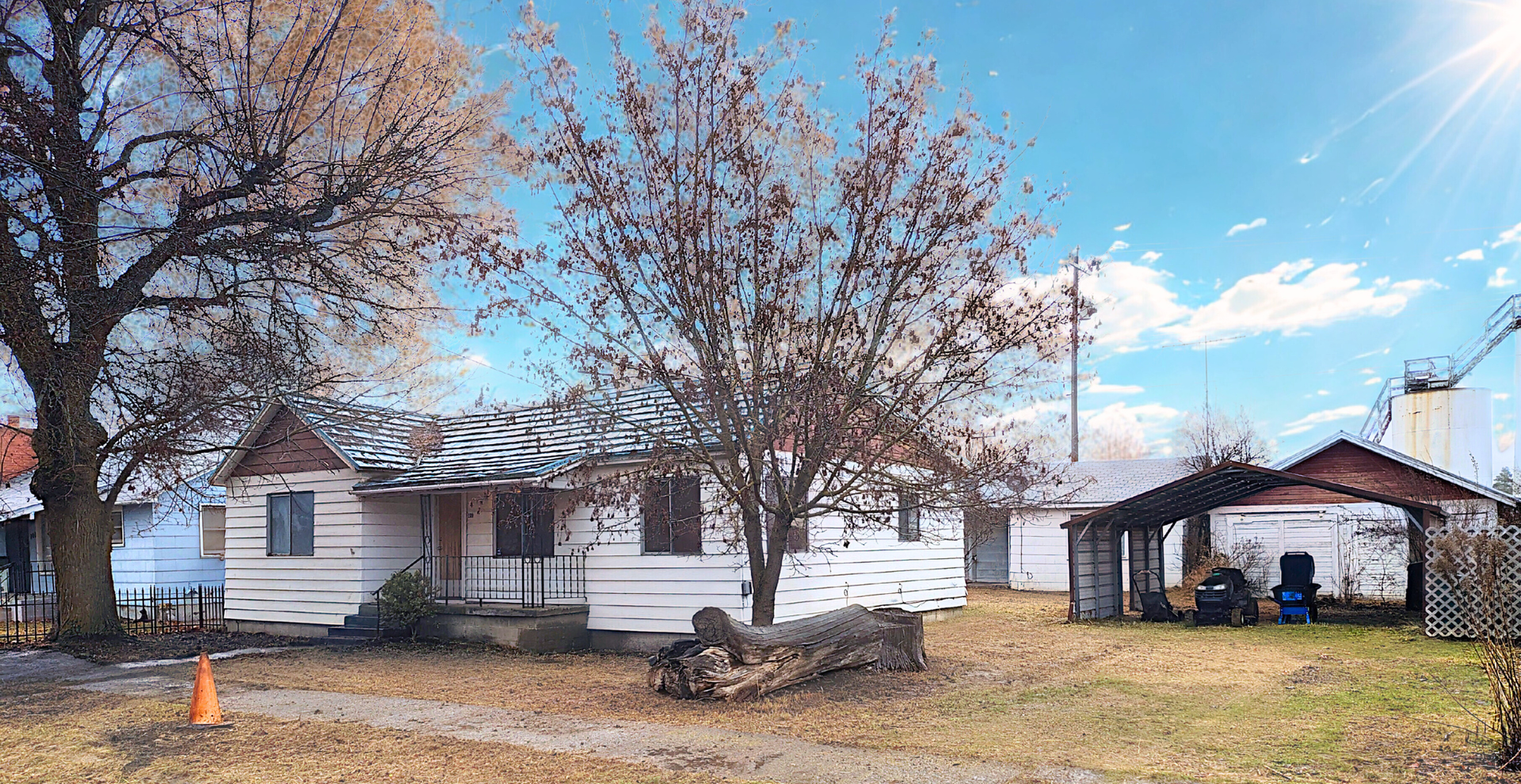 White farmhouse with bare trees and a carport in a grassy yard under a blue sky