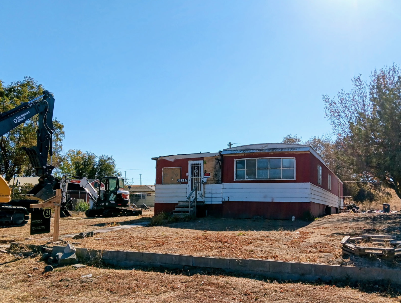 Red mobile home on a dirt lot with construction equipment and stacked lumber under a clear blue sky.