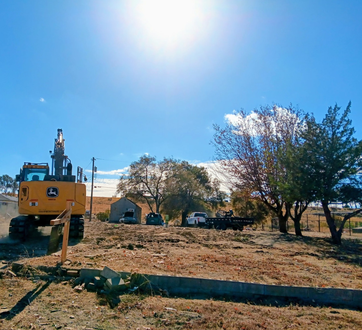 Excavator at a rural worksite under a bright blue sky with trees and a small house in the background