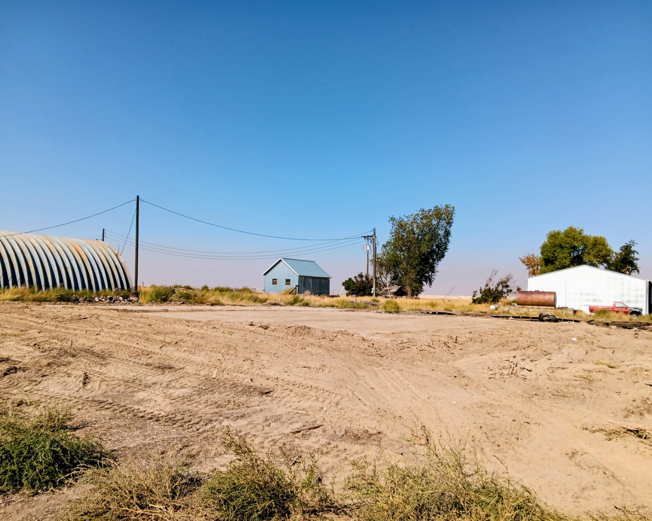Dry rural field with a small house, trees, utility poles, and a blue sky