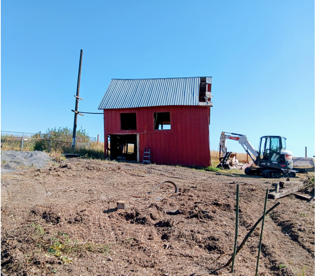 Red barn under construction on a dry field, with an excavator beside it and a blue sky overhead.