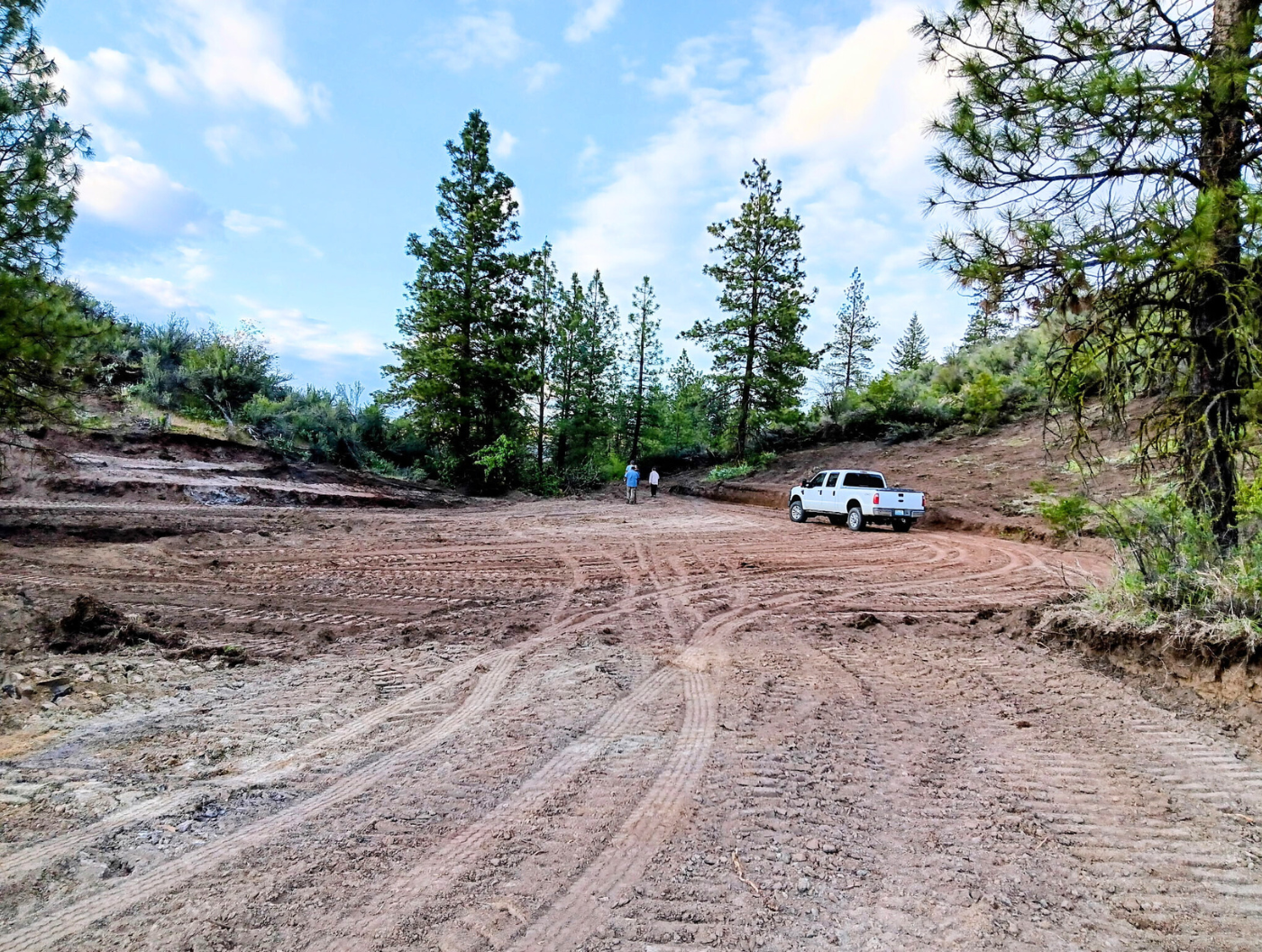 White pickup truck on a muddy dirt road in a wooded clearing under a blue sky.