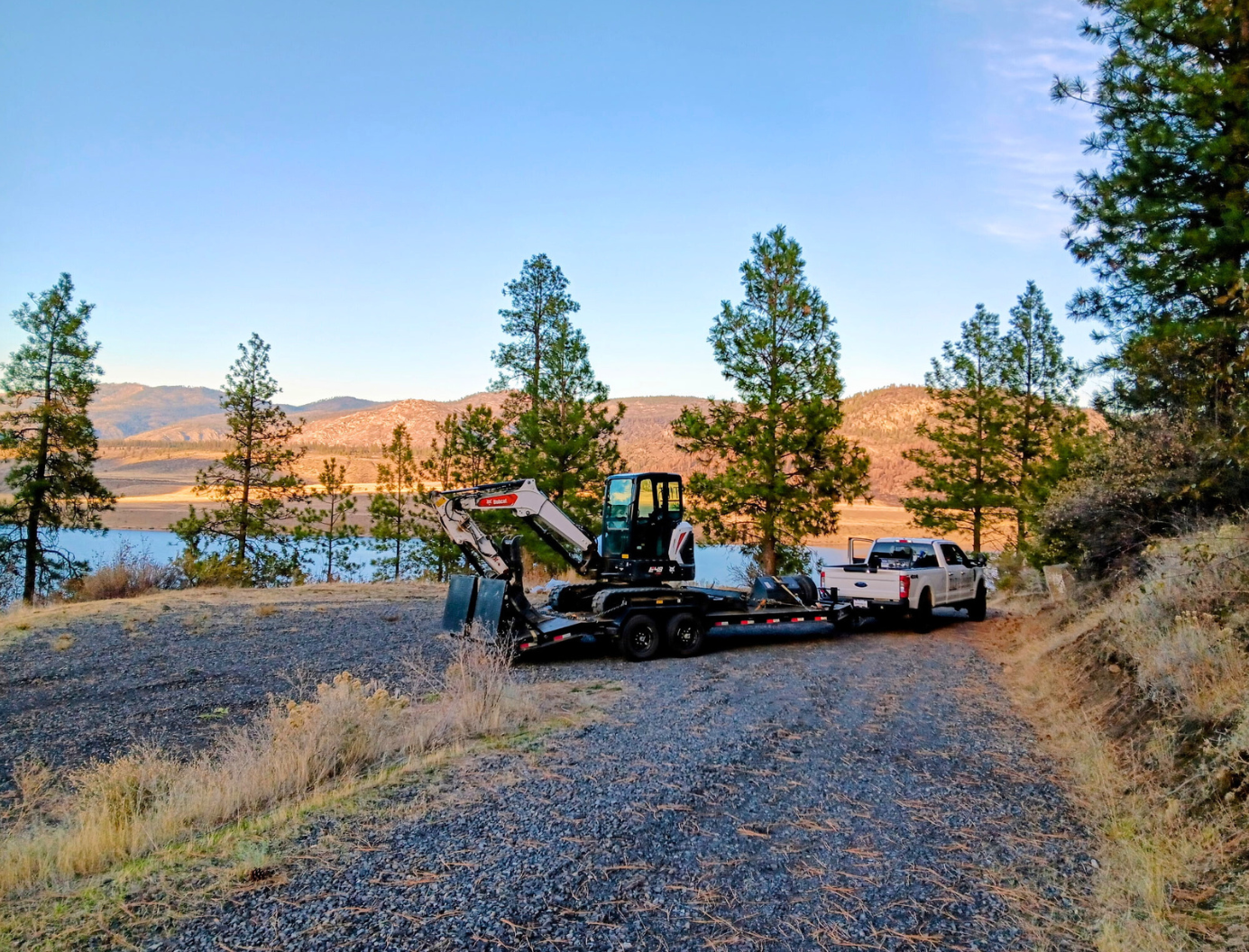 Backhoe and pickup truck on gravel by a lake, with trees and hills at sunset