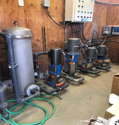 Industrial pump room with five water pumps and tanks along a wood-paneled wall, hoses on the floor.