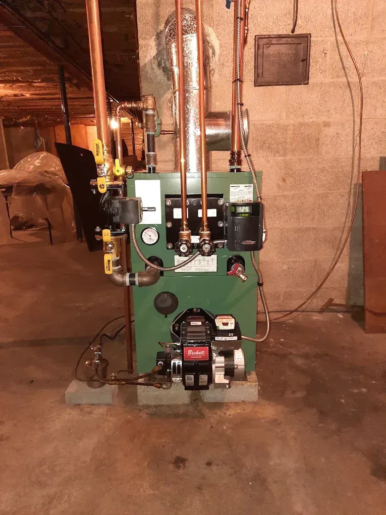 Green boiler/furnace unit in a basement with exposed pipes and two pumps in front.