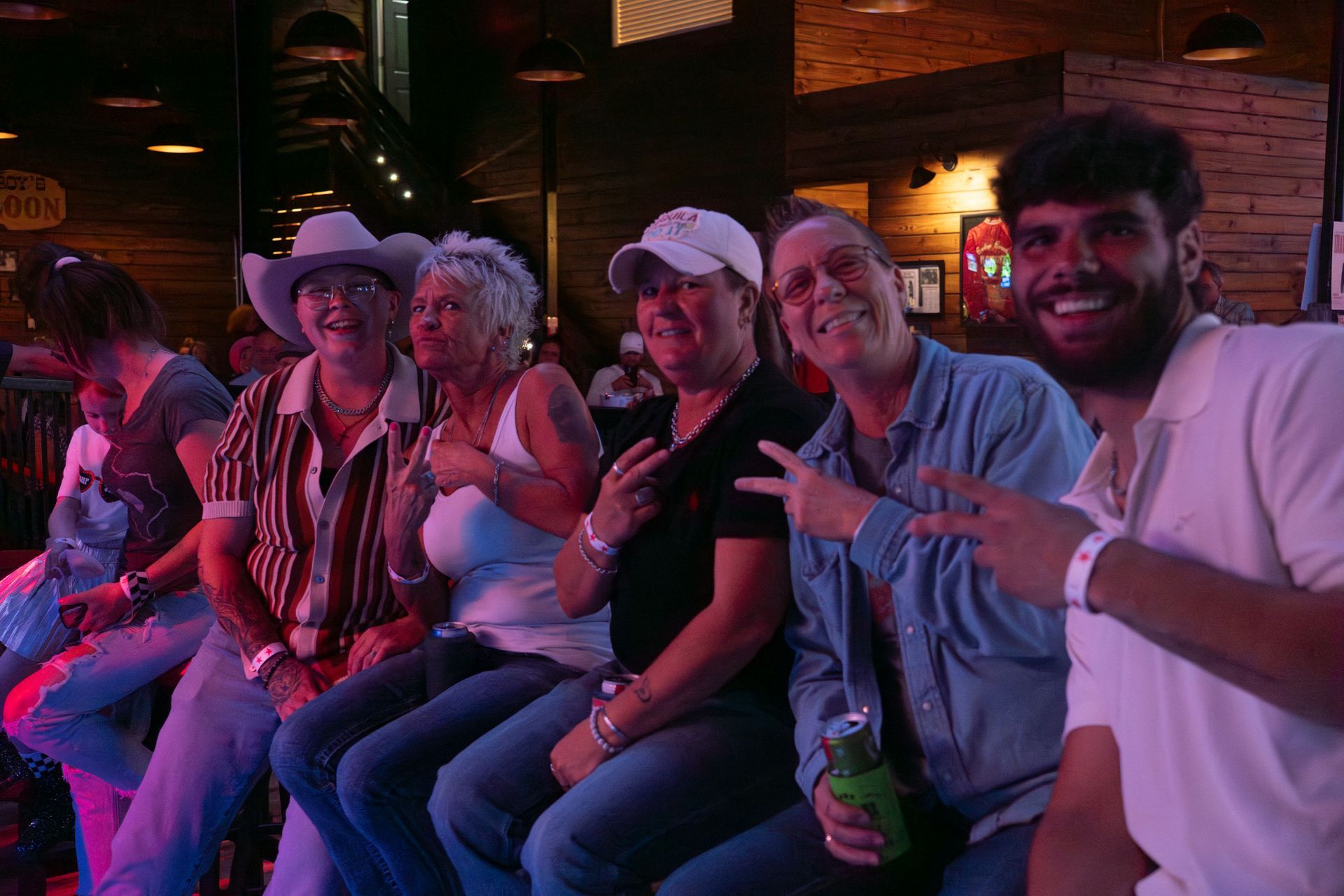 Six smiling friends in cowboy hats pose at a dimly lit venue with neon pink and blue lighting.