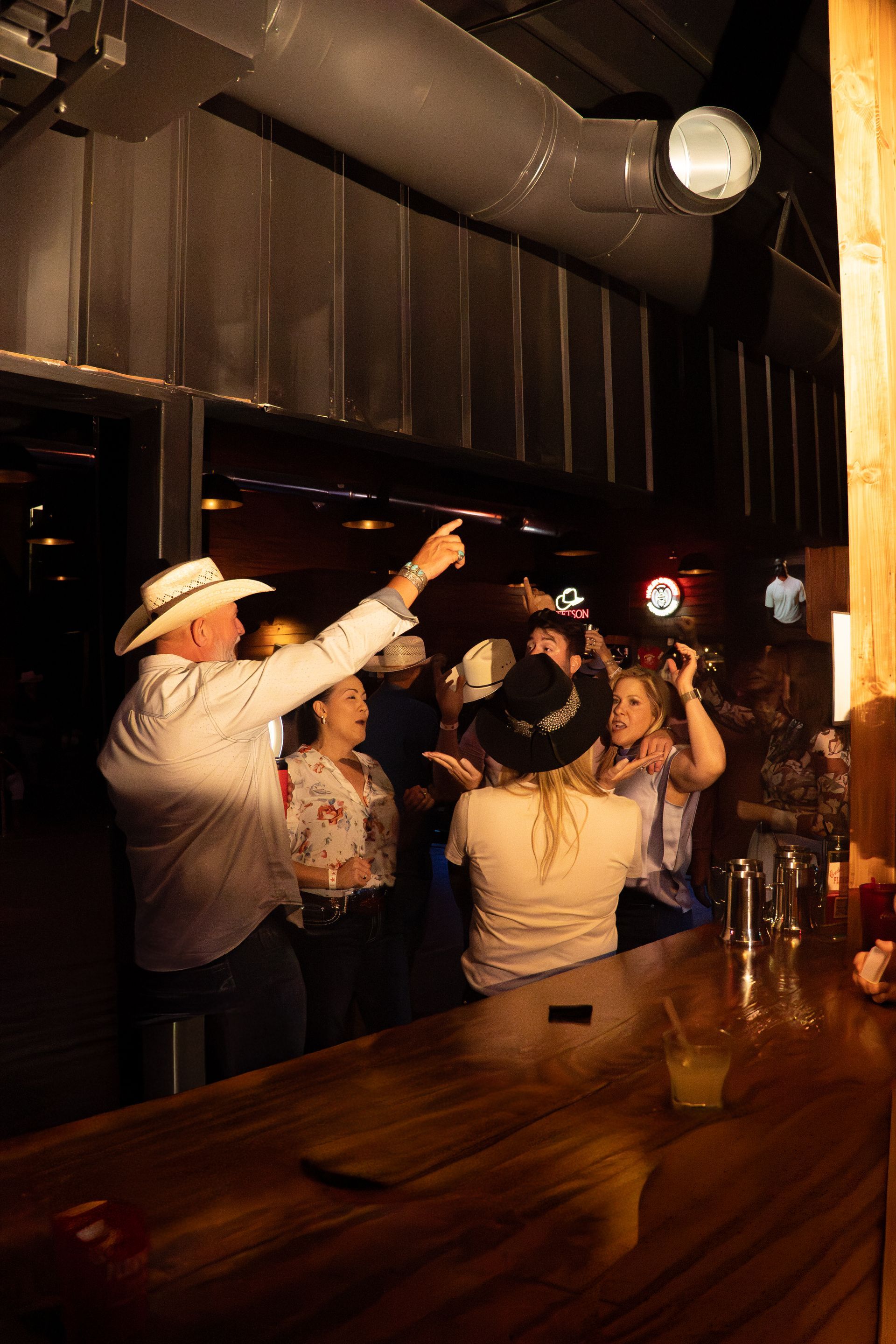 People dancing in a dimly lit bar under warm lights, with a cowboy hat visible at left.